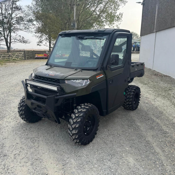 Front three-quarter view of a dark green off-road utility vehicle with large knobby tires and a roof, parked on a gravel lot near a barn.
