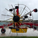 Red and yellow farming implement with long arms and metal tines, resting on a concrete surface under a cloudy sky