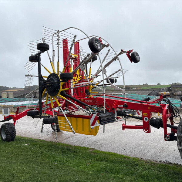 Wide view of a red agricultural implement with multiple tined arms and wheels, parked on a paved area with green grass and cloudy sky behind.
