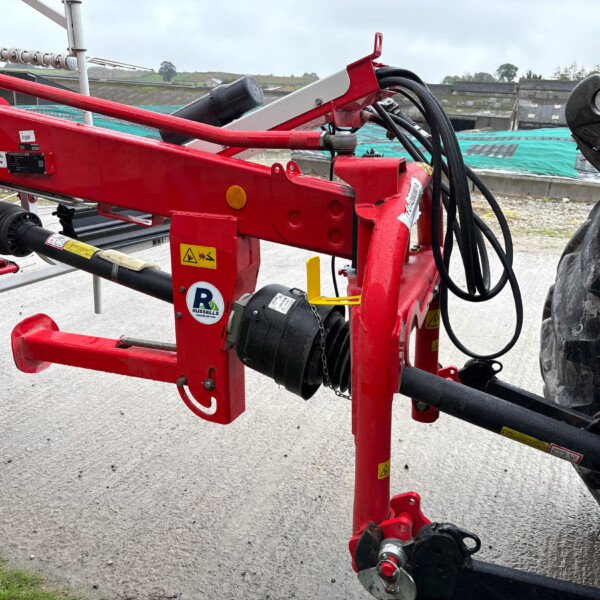 Red agricultural implement frame with hydraulic hoses and a large tractor tire on a construction site backdrop.