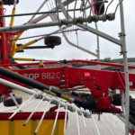 Close-up of a red and yellow construction machine frame with steel bars, hoses, and a wheel, outdoors on a cloudy day.