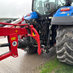 Blue tractor with a red frontend attachment on a gravel path, muddy tires and green grass nearby.