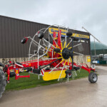 Large red and yellow agricultural mower/tedder with multiple tines parked beside a building outdoors.
