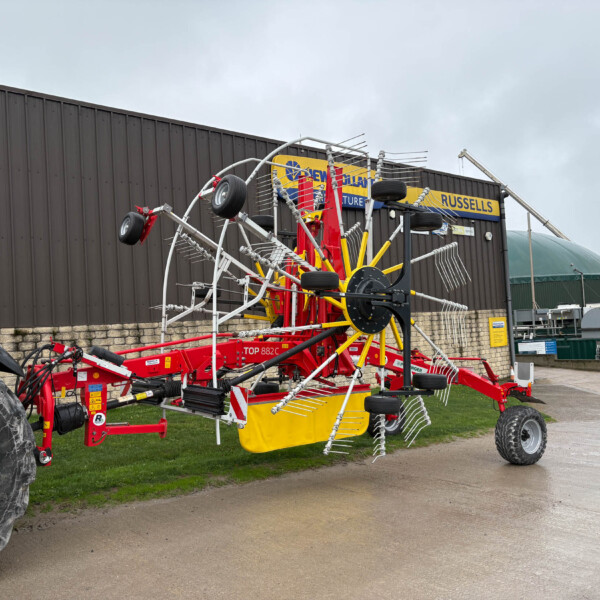 Large red and yellow agricultural mower/tedder with multiple tines parked beside a building outdoors.