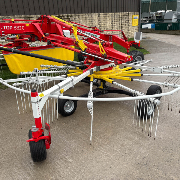 Red agricultural implement with extendable arms and many white tines, yellow guard, on wheels in an outdoor yard near buildings.