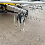 Trailer-mounted agricultural harrow with many vertical tines hanging down, rust-tipped at the tips, on a gravel surface