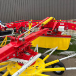 Red and yellow agricultural implement attached to a blue tractor, parked beside a brick wall, with long hydraulic arms and multiple tines extended outward.