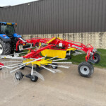 Blue farm tractor with a red/yellow farming implement attached, parked against a tall gray metal building and a stone base wall.