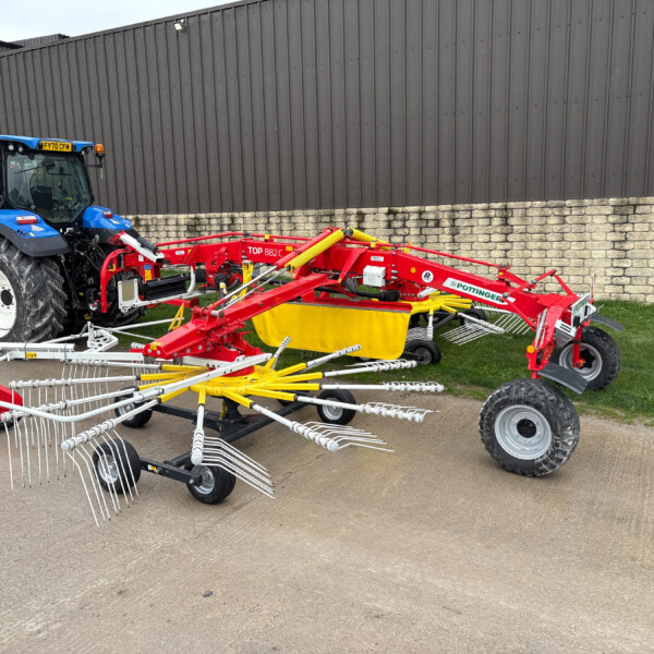 Blue farm tractor with a red/yellow farming implement attached, parked against a tall gray metal building and a stone base wall.