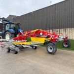 Blue tractor with a red multi-rake implement attached, parked beside a dark corrugated metal building.