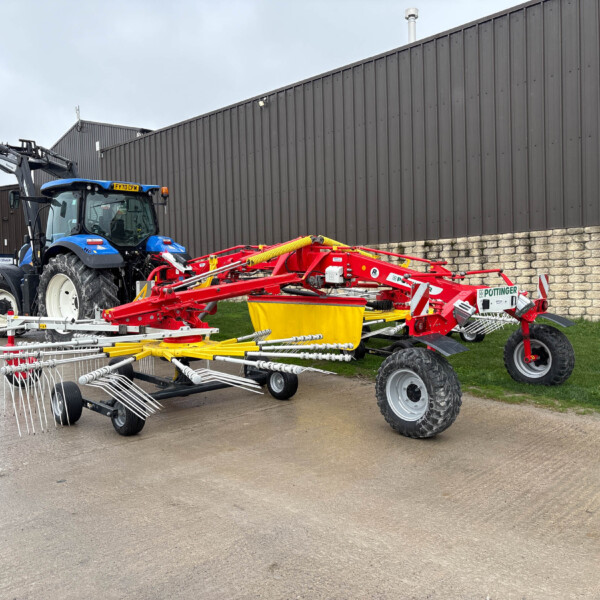 Blue tractor with a red multi-rake implement attached, parked beside a dark corrugated metal building.