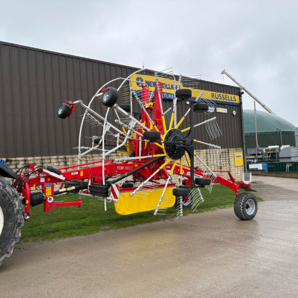 Red multi-row agricultural implement with yellow spokes and white tines on a trailer, displayed in a yard beside a dark industrial building with a Russell sign.