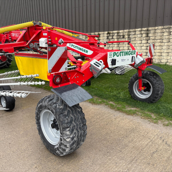 Red Pöttinger agricultural implement attached to a blue tractor, parked beside a stone wall with a yellow hose on top and metal tines visible in the foreground.