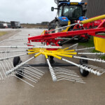 Blue tractor with large red implement in a yard, long yellow and white tines spread outward, waiting equipment in background.