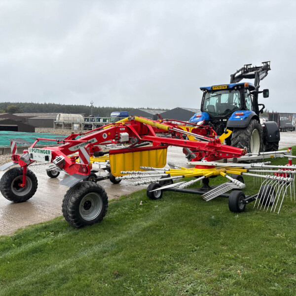 Blue tractor with red farming implements in a yard near farm buildings on a cloudy day.