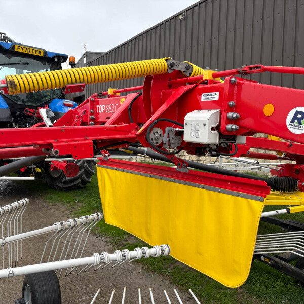 Red trailer-mounted farming implement attached to a blue tractor, featuring a yellow coiled hose and multiple tines, beside a corrugated metal wall in a farm lot.