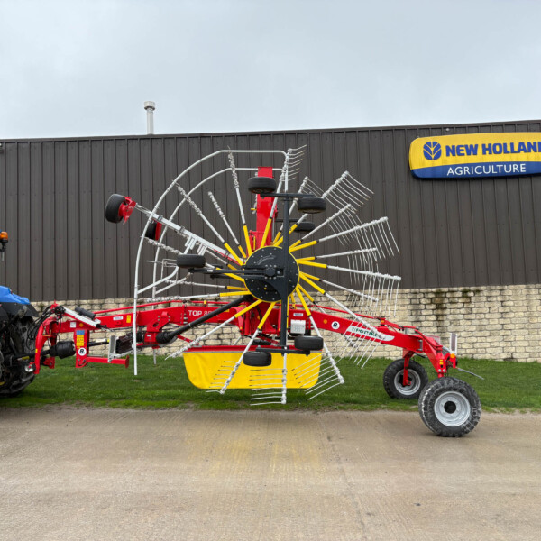 Blue tractor parked beside a red-and-silver rotary rake implement, in front of a dark metal building with a New Holland Agriculture sign outside.