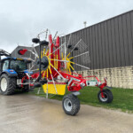 Blue tractor hitched to a large red and yellow hay rake with many metal tines, parked beside a dark industrial building.