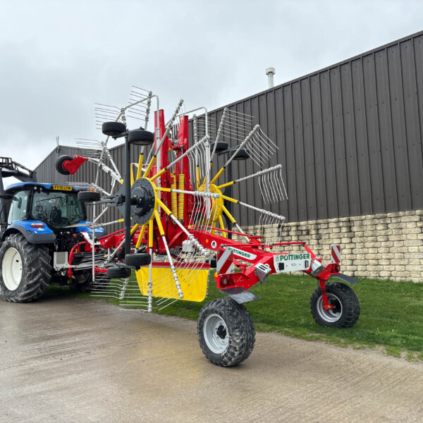 Blue tractor hitched to a large red and yellow hay rake with many metal tines, parked beside a dark industrial building.