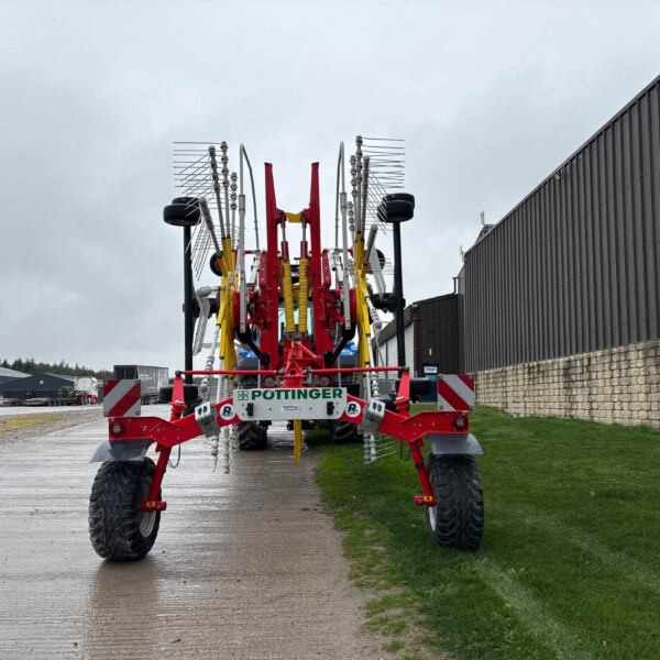 Front view of a large red and yellow agricultural spray machine with folded spray booms, parked on a wet road beside a corrugated metal building.