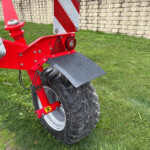 Rear view of a red bike trailer with a large mud-covered tire, reflector, and a safety sign, on grass near a stone wall.