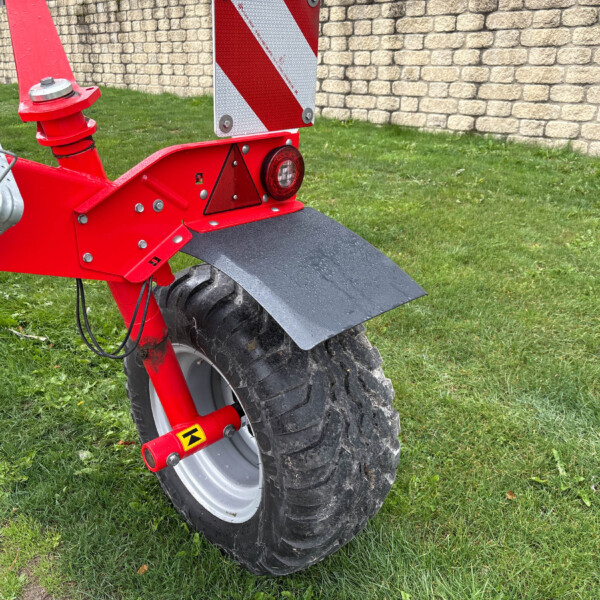 Rear view of a red bike trailer with a large mud-covered tire, reflector, and a safety sign, on grass near a stone wall.