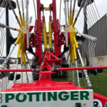 Close-up of a red and yellow agricultural harvester with long curved tines and yellow springs, outdoors in a farm yard. Brand 'POTTINGER' visible on the equipment sign.