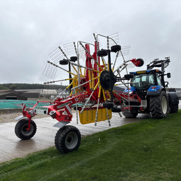 Tractor pulling a large red and yellow cultivator with numerous tines and wheels in a farmyard under a cloudy sky.