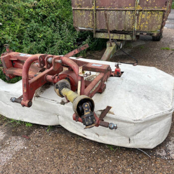 Old rusty red farming equipment resting on a white bag, with dense greenery behind and a small brown-and-white dog on the gravel path to the right.