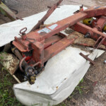 Old rusty agricultural implement on top of a stained white mattress outdoors, with coiled yellow hoses and overgrown grass nearby.