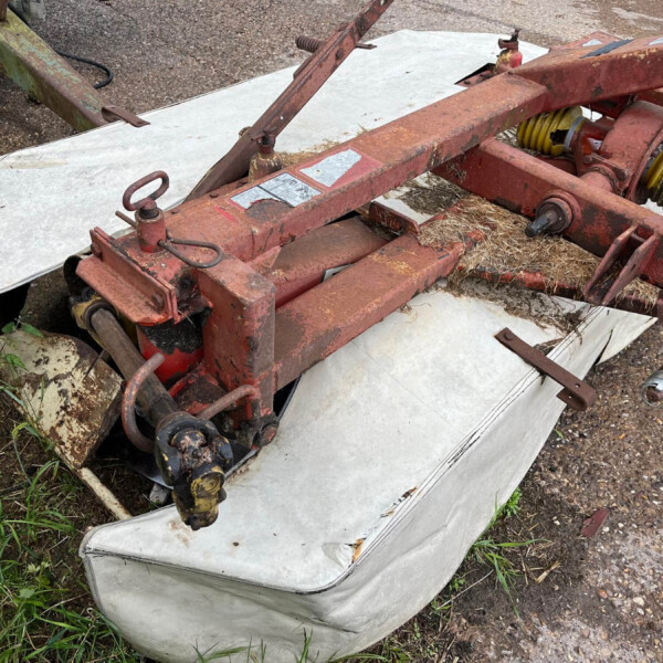 Old rusty agricultural implement on top of a stained white mattress outdoors, with coiled yellow hoses and overgrown grass nearby.