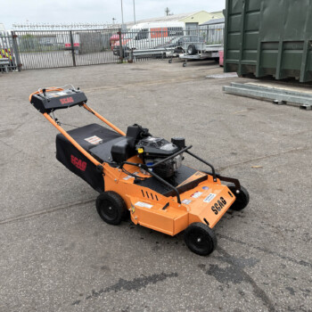 Orange SCAG turf mower in a paved yard with metal fencing and a large green dumpster on the right, parked near cargo trailers and pallets.