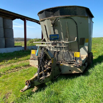 Large agricultural machine with cables and a digital display, outdoors on a grassy field beside a shed and silo stacks under a blue sky.
