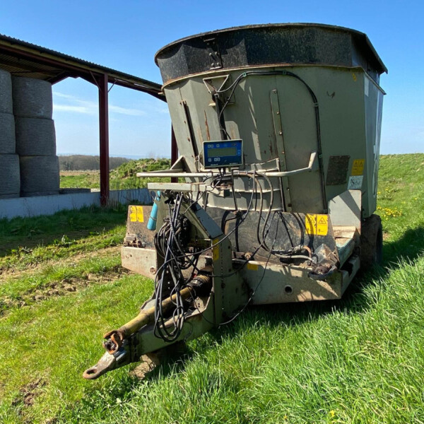 Large agricultural machine with cables and a digital display, outdoors on a grassy field beside a shed and silo stacks under a blue sky.