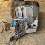 Old hay baler with hoses and a metal frame sits among stacked straw bales in a barn.