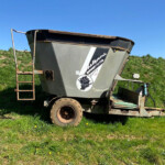 Large gray grain cart on a trailer parked in a grassy field under a blue sky, with a rusty metal ladder on the left.