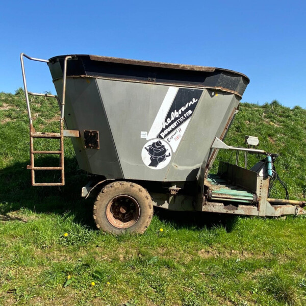 Large gray grain cart on a trailer parked in a grassy field under a blue sky, with a rusty metal ladder on the left.
