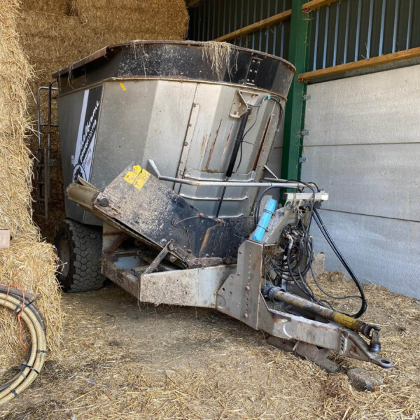 Farm harvester sits tipped on its side next to large stacks of straw bales inside a shed.