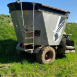 Large gray agricultural trailer tilted on muddy wheels in a grassy field with a clear blue sky, attached to a tractor edge.