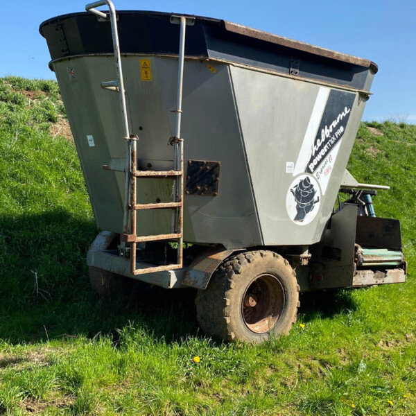 Large gray agricultural trailer tilted on muddy wheels in a grassy field with a clear blue sky, attached to a tractor edge.
