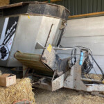 Industrial hay harvester/auger in a shed, surrounded by tightly packed straw bales and scattered cardboard boxes.