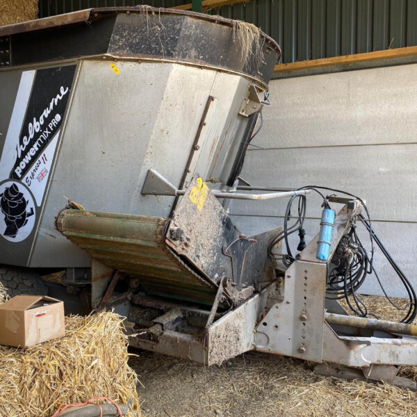 Industrial hay harvester/auger in a shed, surrounded by tightly packed straw bales and scattered cardboard boxes.
