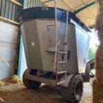 Large agricultural baler or forage wagon inside a barn, surrounded by dry straw bales and wooden beams.
