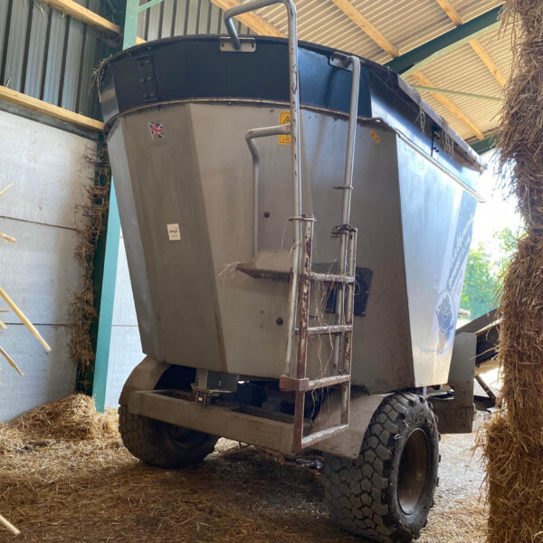 Large agricultural baler or forage wagon inside a barn, surrounded by dry straw bales and wooden beams.