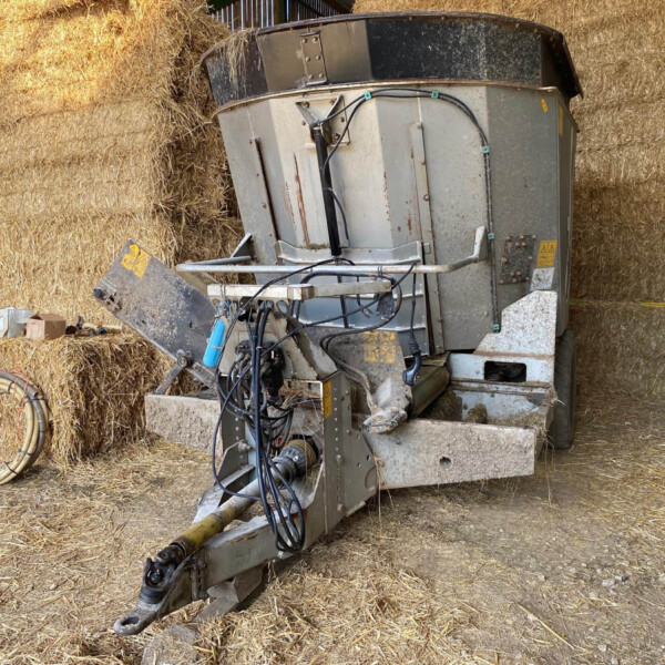 Old hay baler with hoses and a metal frame sits among stacked straw bales in a barn.
