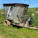 Tilted gray farm grain cart trailer resting on its side in a grassy field under a clear blue sky, with rusted metal and hoses visible.