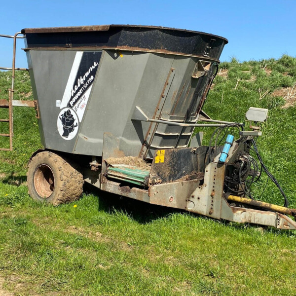 Tilted gray farm grain cart trailer resting on its side in a grassy field under a clear blue sky, with rusted metal and hoses visible.