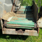 Rusty farm conveyor with green slats and soil on the belt, surrounded by tall grass and weeds.