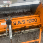 Close-up of an orange industrial machine with a perforated gray metal belt, showing rust and dirt; a sticker reads 'golf & turf machinery' on top.