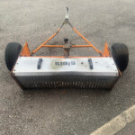 Top-down view of an orange metal trailer frame with two large black wheels on a paved surface.